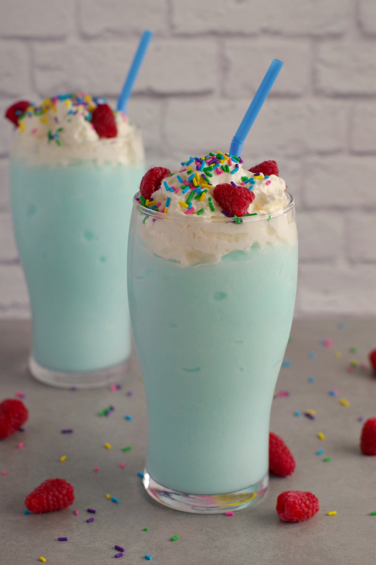 2 blue raspberry slushie drinks on grey counter with sprinkles and raspberries
