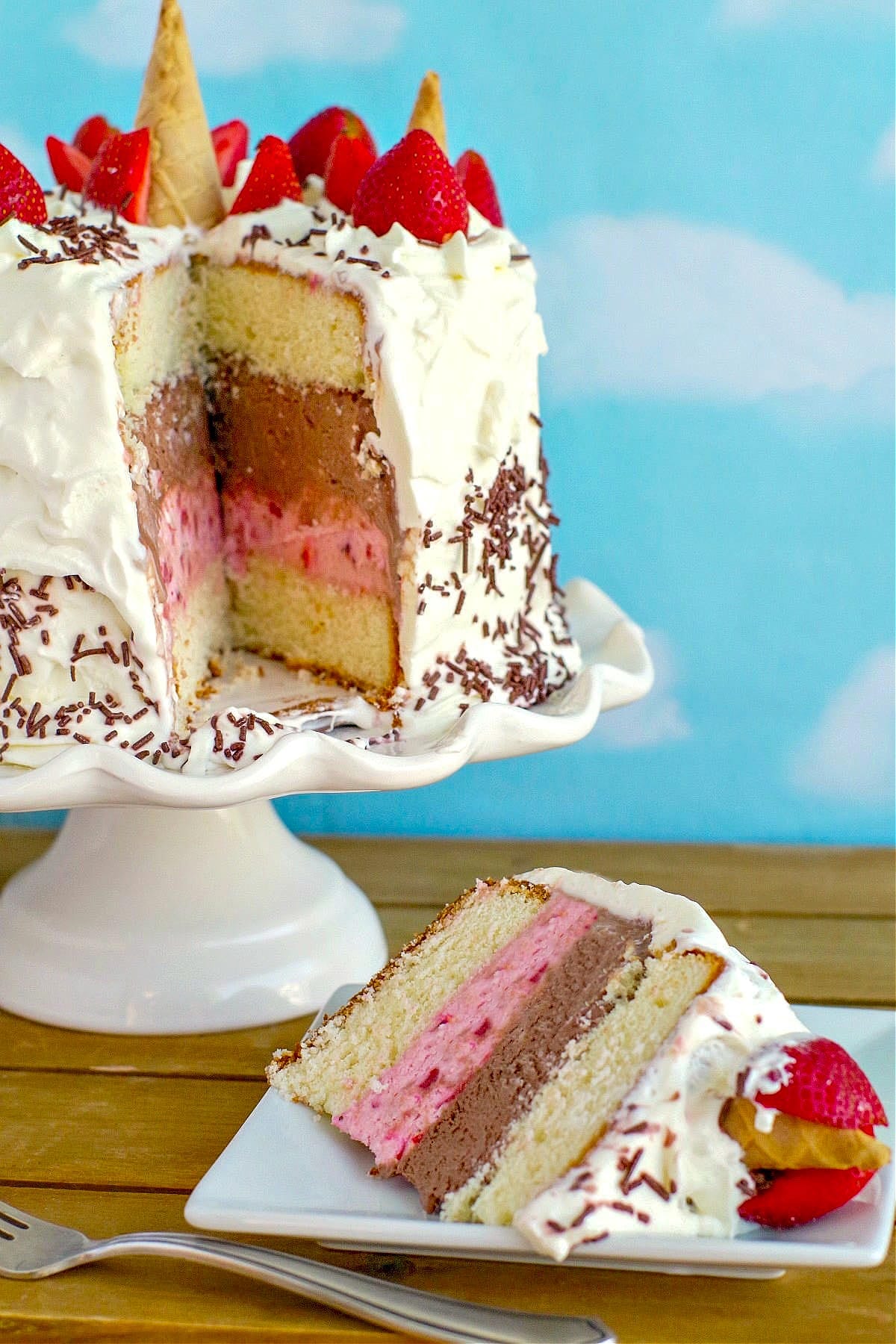 a slice of Neapolitan Ice Cream cakes on a white plate, with whole cake on white cake stand in the background