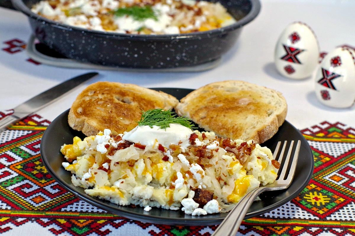 Scrambled Ukrainian style eggs on a black plate in foreground with pan of fried Ukrainian style eggs in background