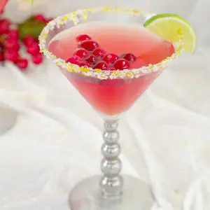 Cranberry Cosmopolitan Mocktail with cranberries floating in drink, wedge of lime and sparkly gold sugar with cranberries in the background, on white material