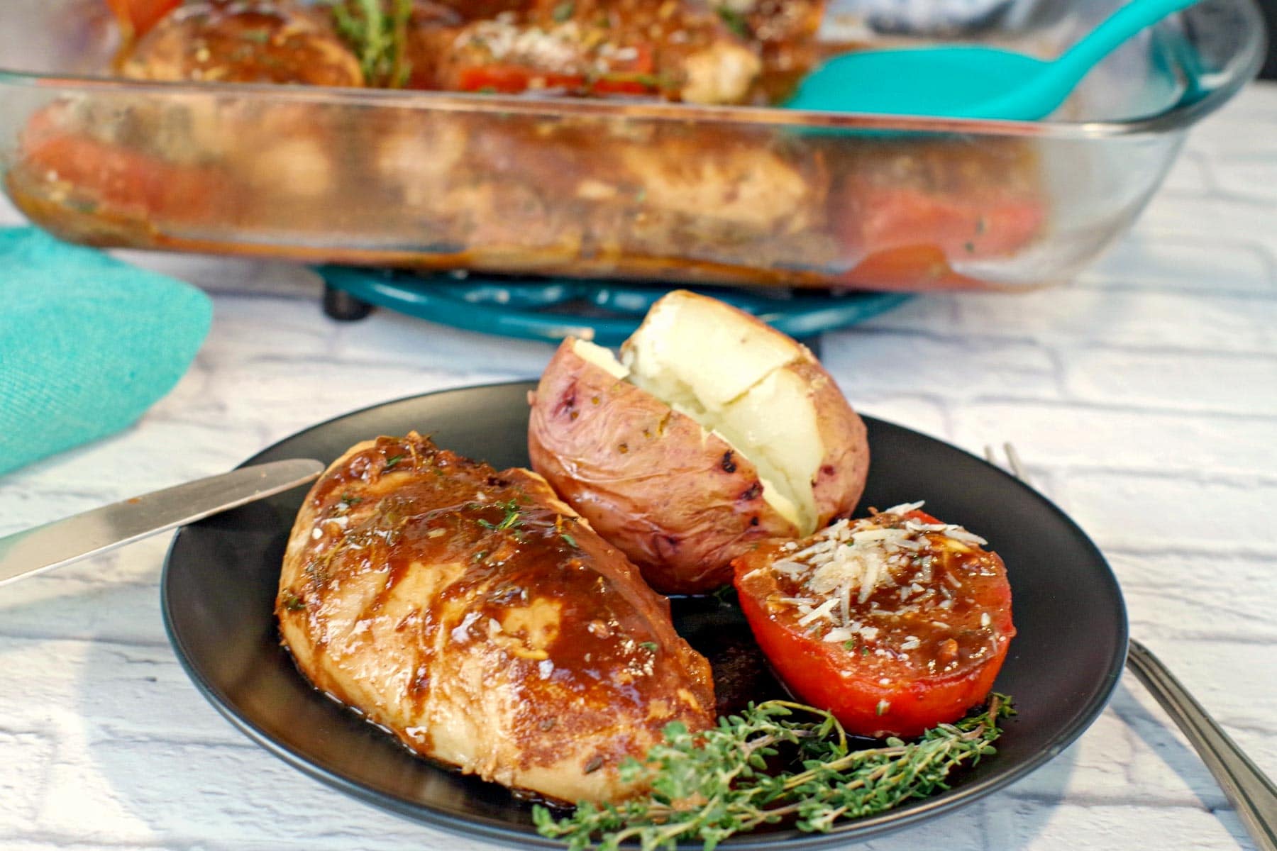 Balsamic chicken and tomatoes with baked potato on a black plate with a glass container of balsamic chicken in background