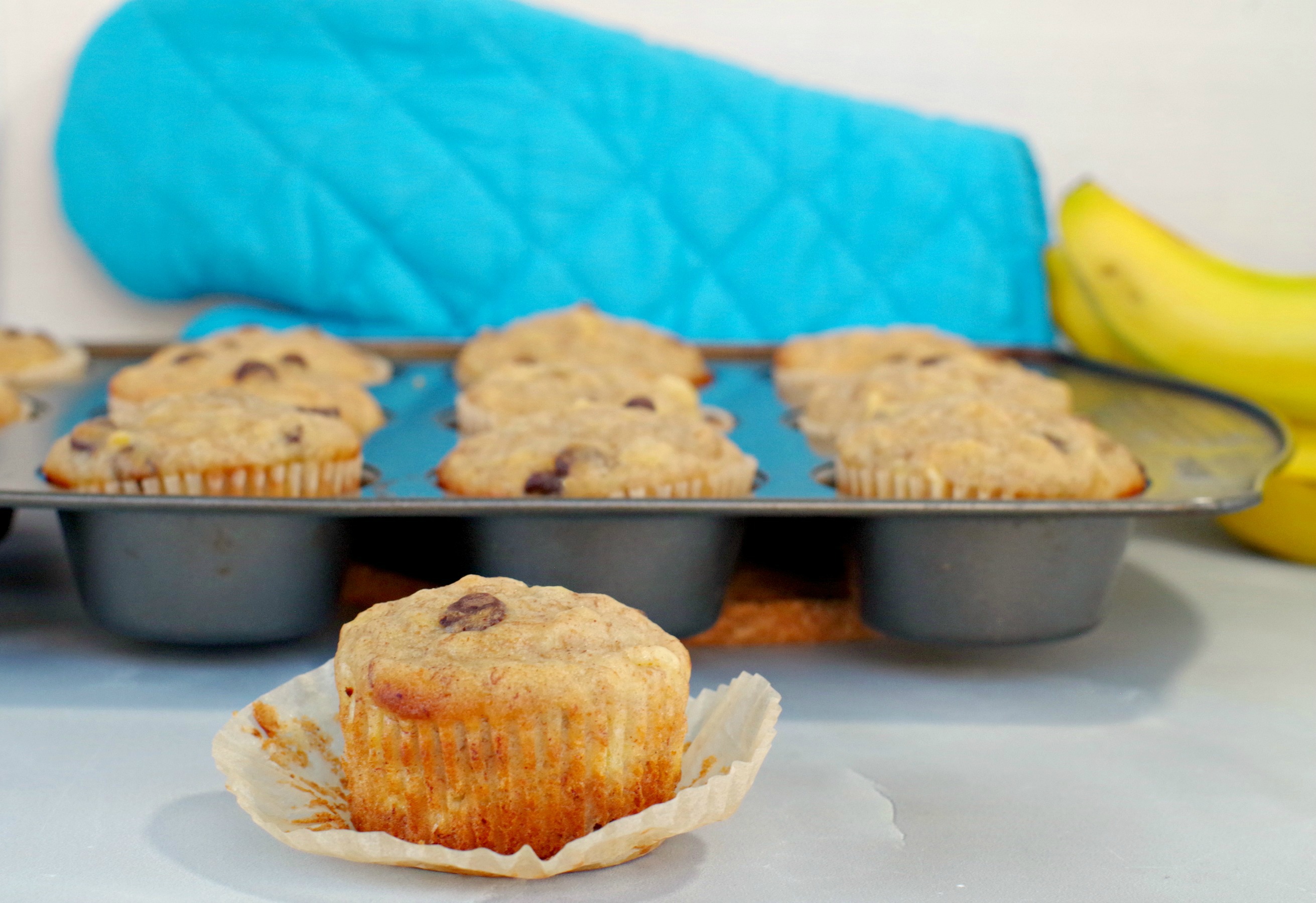 Healthy Banana Chocolate Chip muffins with pineapple in a muffin tin in background with muffin (with liner off) in front