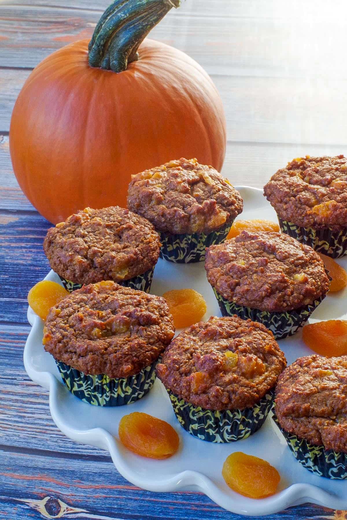 healthy bran pumpkin muffins on a white cake platter with dried apricots and a pumpkin in the background