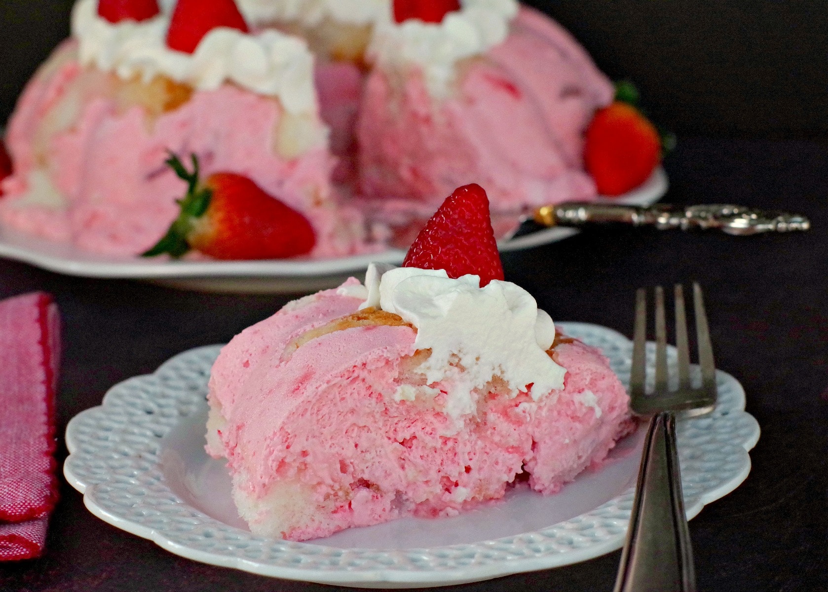 Piece of strawberry jello angel food cake on white plate with whole cake in background