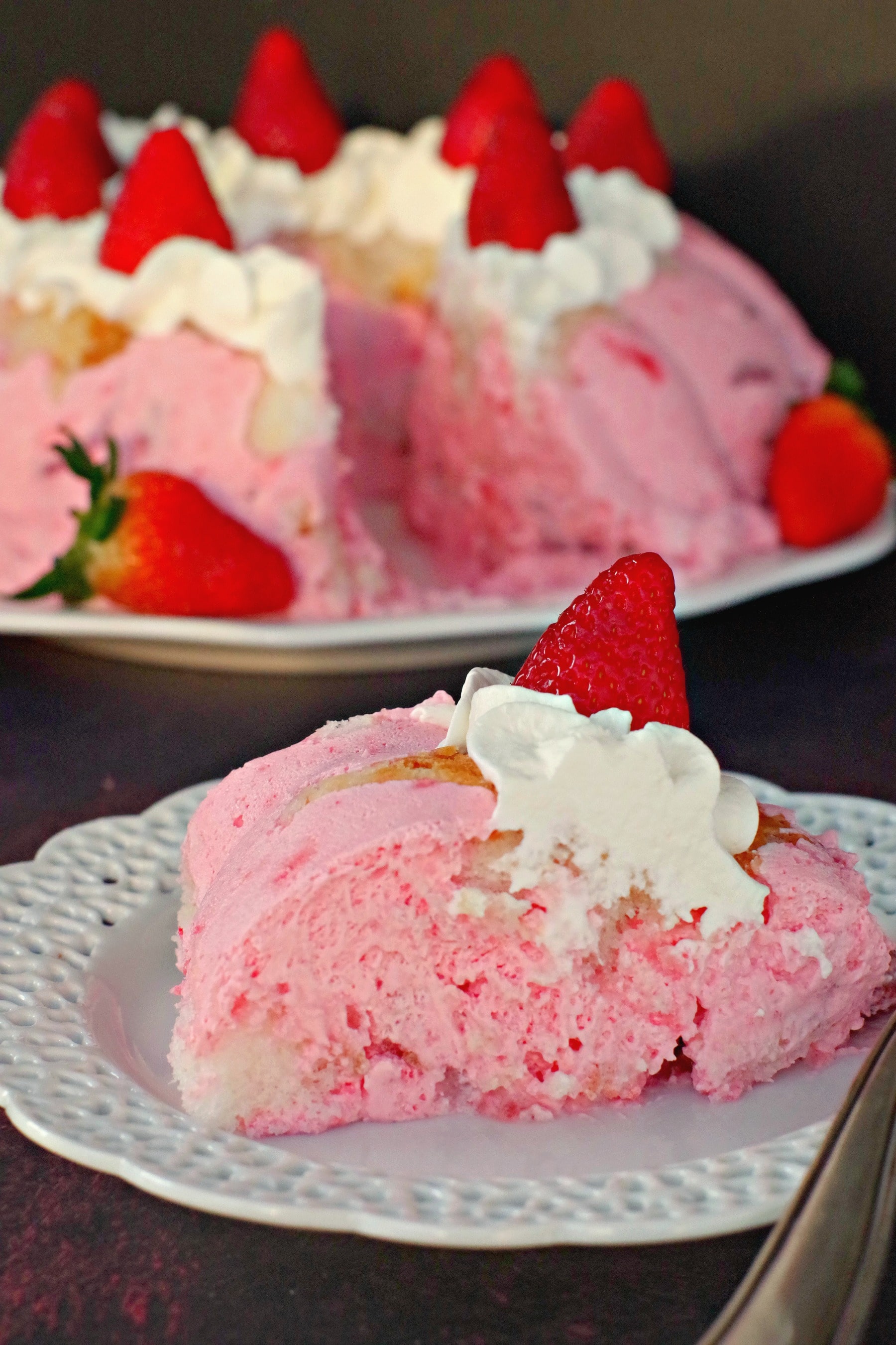 Piece of strawberry jello angel food cake on white plate with whole cake in background