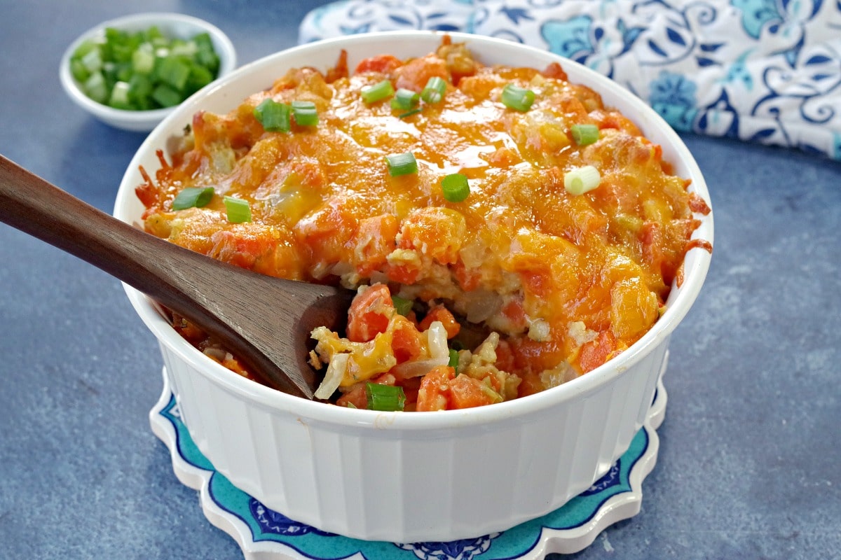 carrot casserole in white casserole dish with a brown wooden spoon scooping casserole (on blue background) with dish of green onions and oven mitts in the background