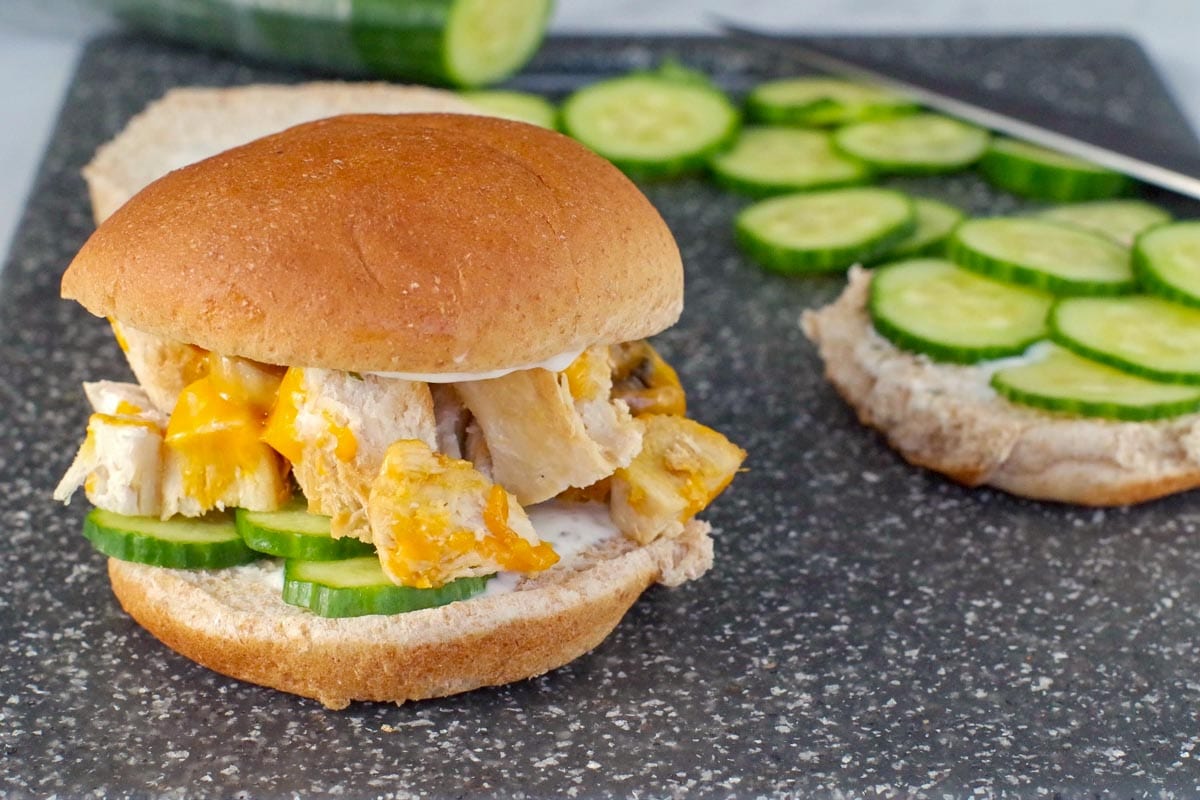 Chicken burger on a cutting board with a ½ bun with cucumber and more cucumbers in the background