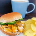 Chicken breast burger on a white plate with potato chips and a blue cup of coffee in the background.
