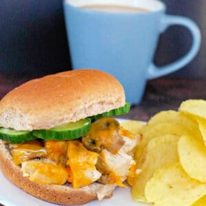 Chicken breast burger on a white plate with potato chips and a blue cup of coffee in the background.