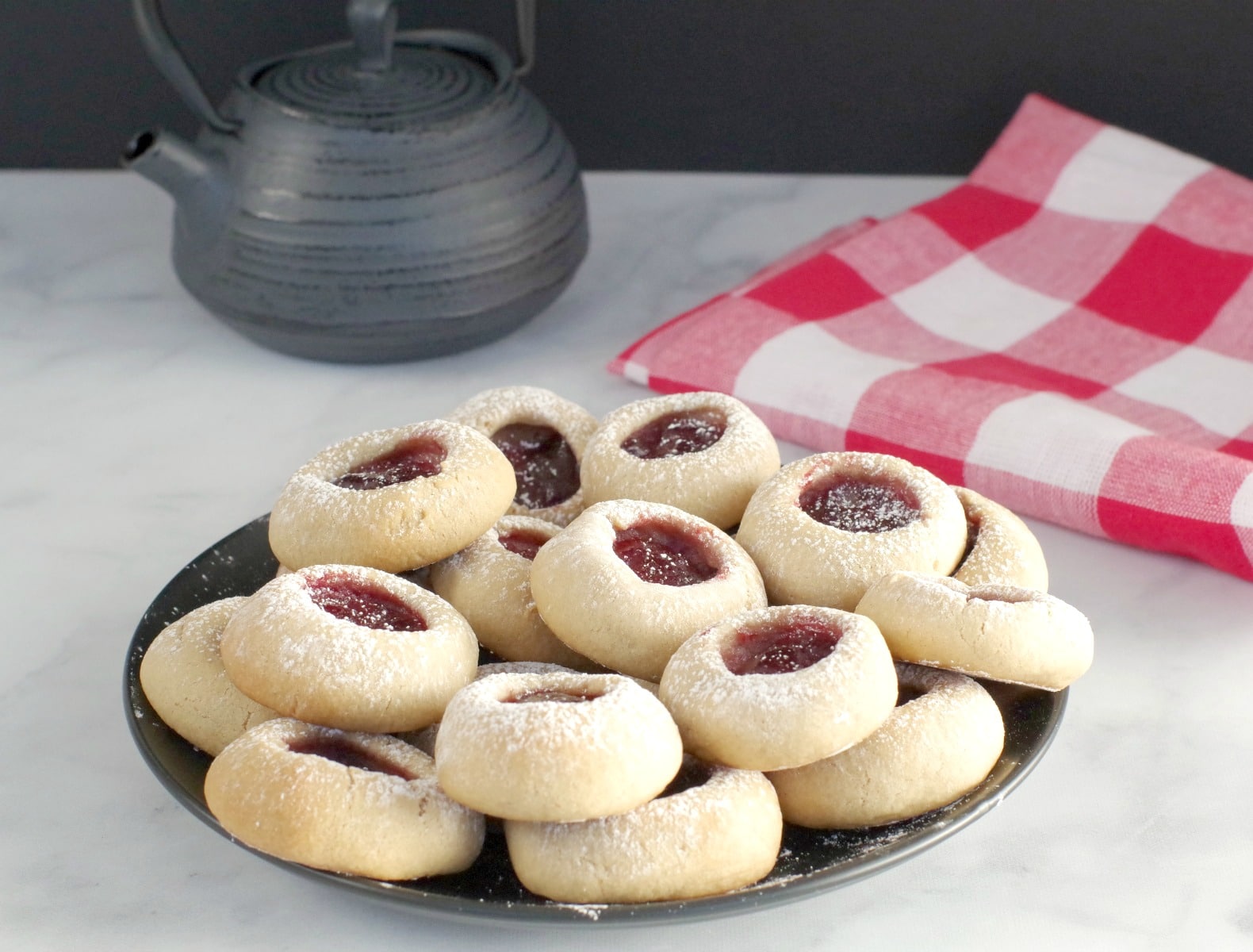 raspberry jam thumbprint cookies on a black plate with black tea pot and red checkered tea towel in the background