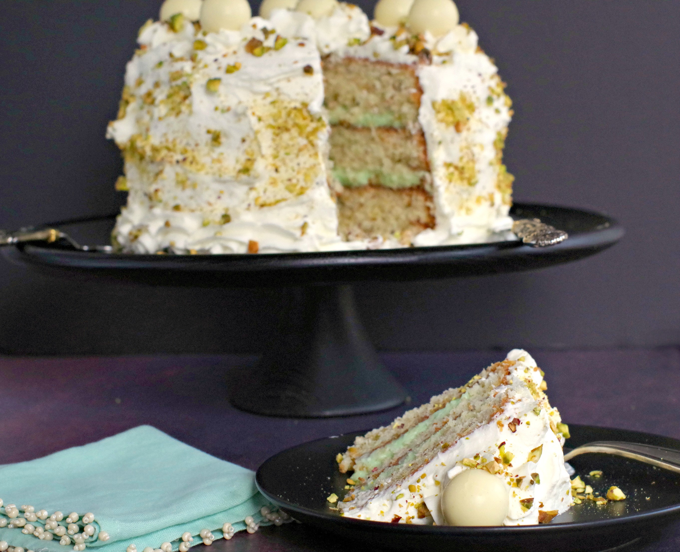 Pistachio White Chocolate Cake on black tray in background with piece of cake in the front