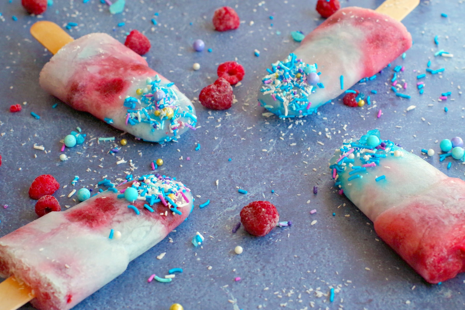 4 blue raspberry popsicles on blue surface with coconut, frozen raspberries and sprinkles all around