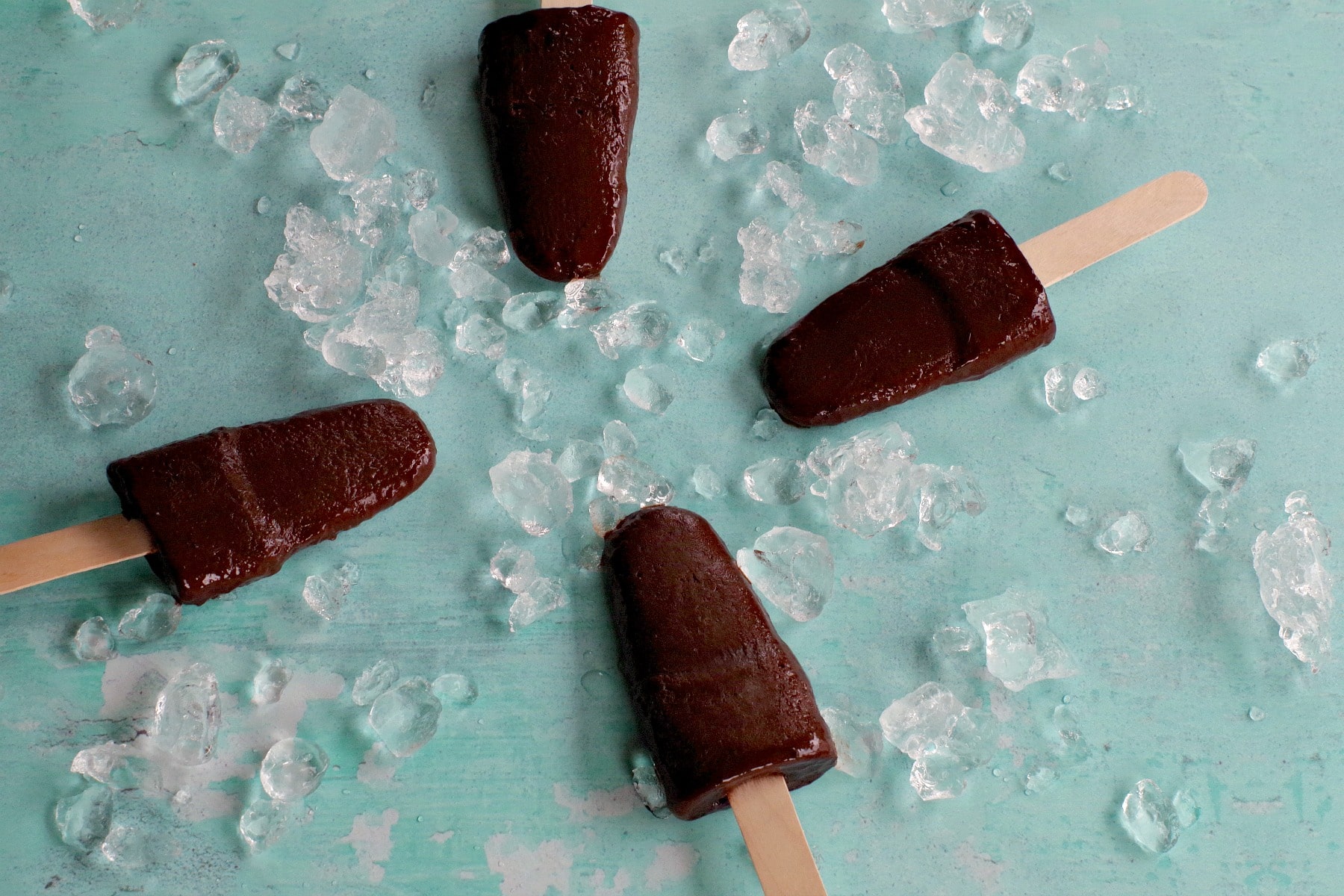 4 chocolate and banana popsicles with ice, laying flat on aqua blue surface