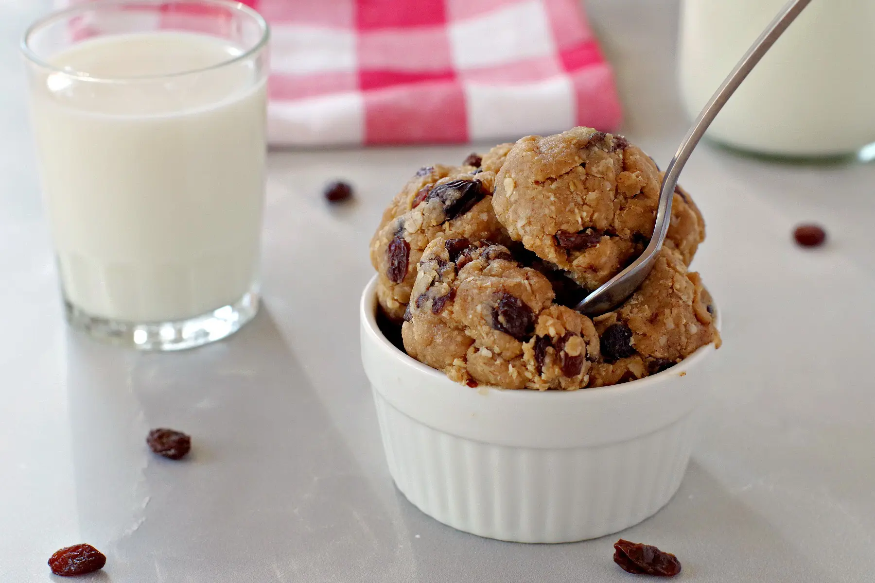 oatmeal raisin edible cookie dough in white bowl with spoon and glass of milk in the background