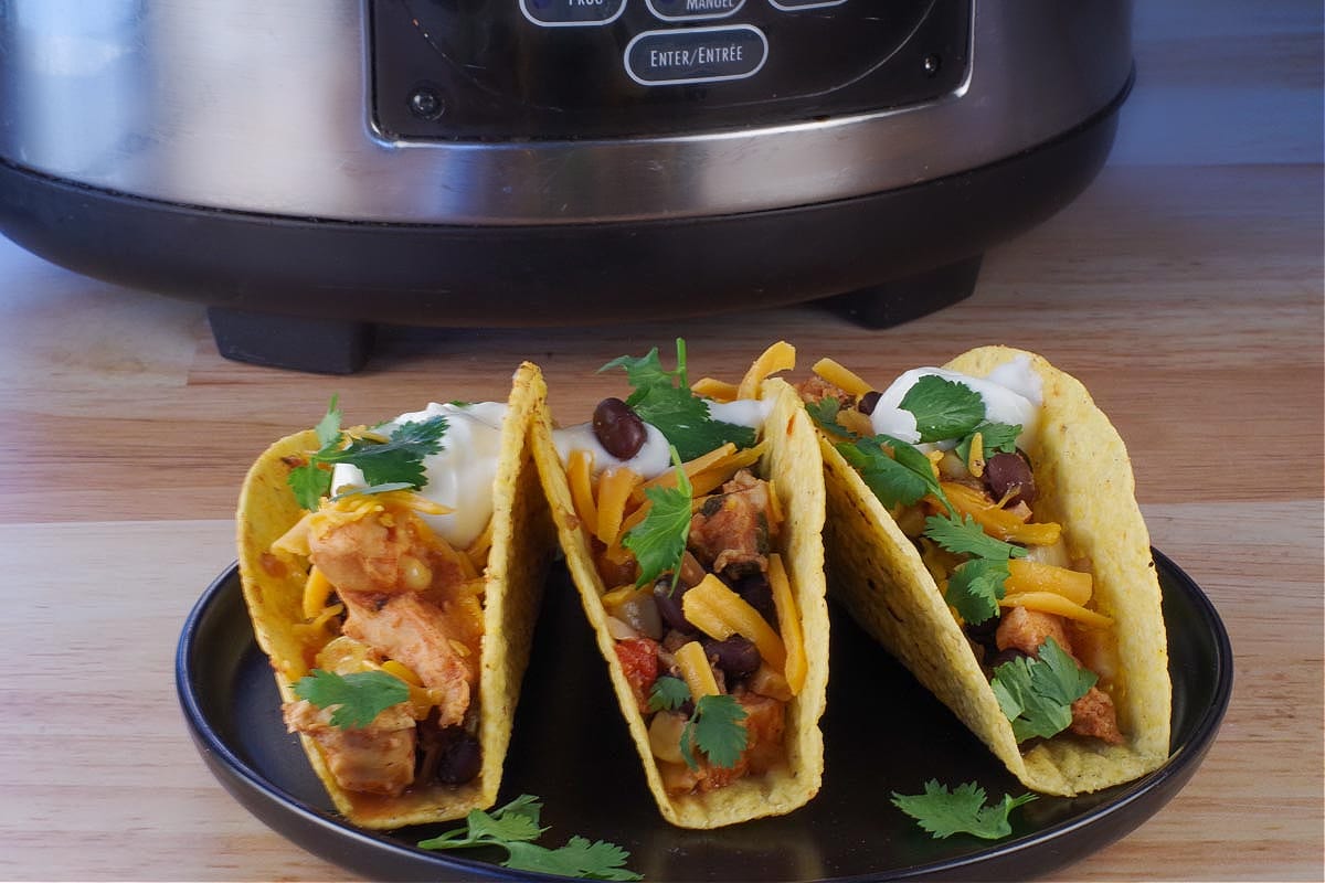 3 Mexican chicken tacos on a black plate with a slow cooker in the background