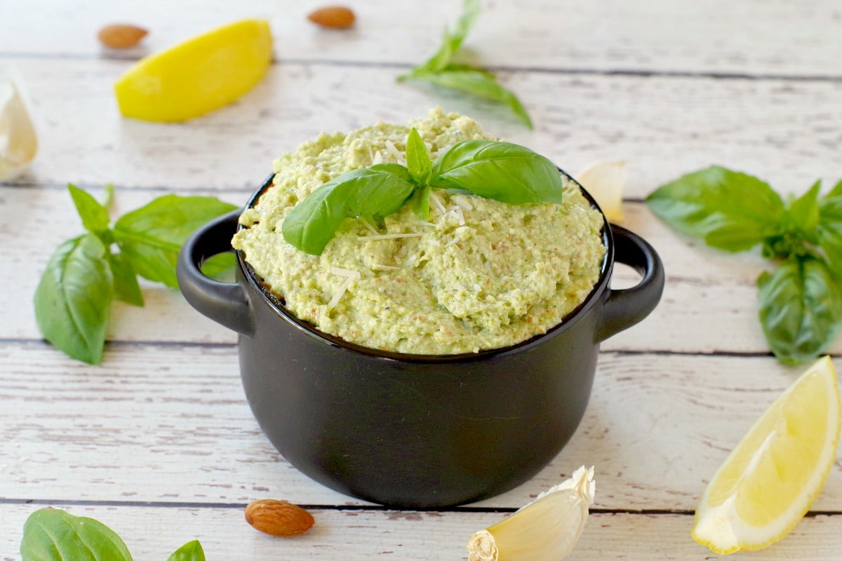 basil pesto with almond in a black dish with almonds, basil, garlic and lemon surrounding the dish