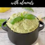 pin with text at the top and photo of basil pesto with almond in a black dish with almonds, basil, garlic and lemon surrounding the dish