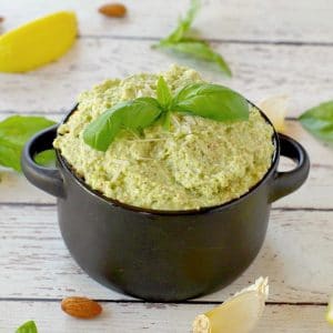 basil pesto with almond in a black dish with almonds, basil, garlic and lemon surrounding the dish