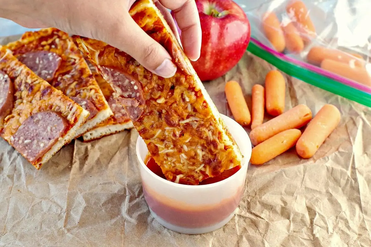 Pizza dipper being dipped into pizza sauce in a small plastic container on a crumpled brown paper bag with and apple and baby carrots in the background.