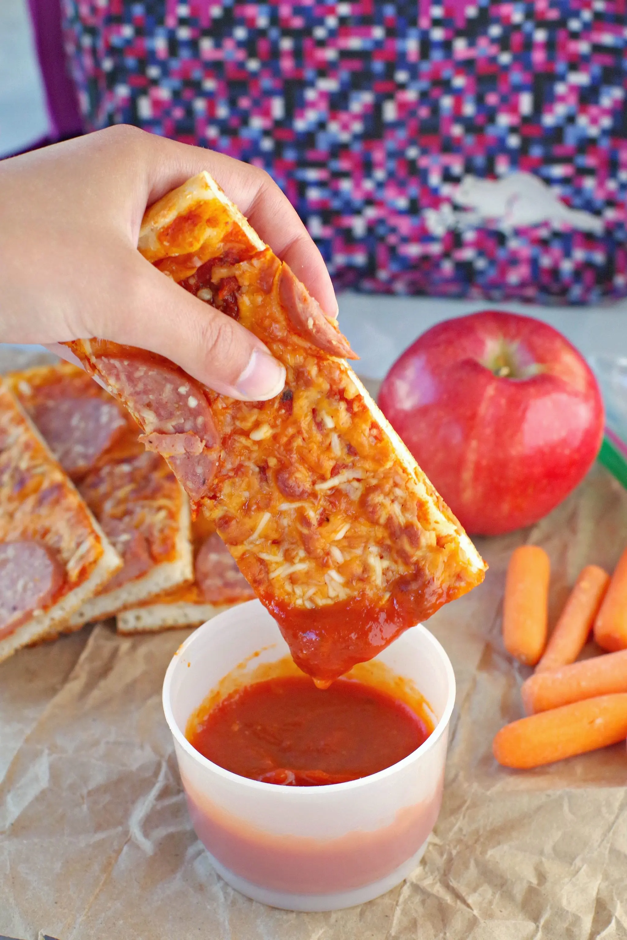 pizza dipper being dipped into pizza sauce with pieces, an apple, carrots and lunch bag in background.