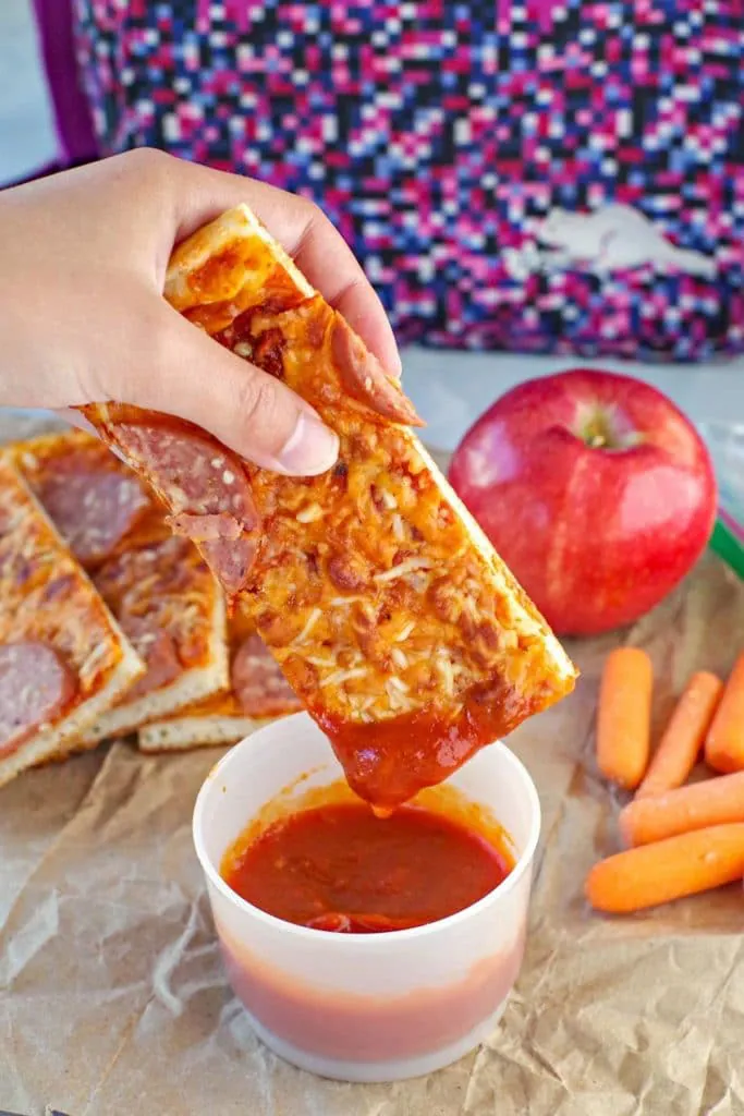 pizza dipper being dipped into pizza sauce with pieces, an apple, carrots and lunch bag in background.