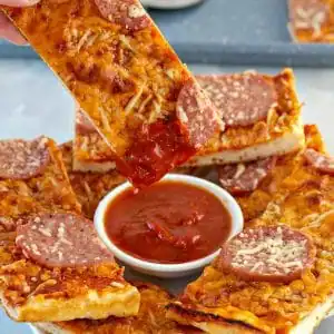 Pizza dipper being dipped into pizza sauce with pieces of pieces around it on white plate. White ranch dip on plate as well.