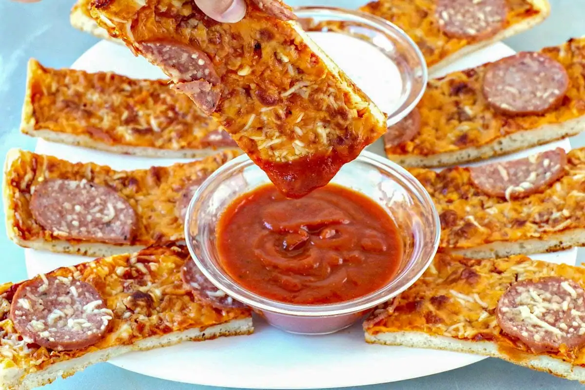 Pizza dipper being dipped into pizza sauce with pieces of pieces around it on white plate. White ranch dip on plate as well.