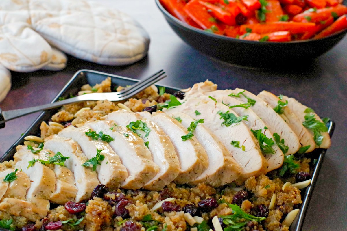 sliced slow cooker turkey breast on bed of cranberry quinoa dressing (on black platter) with black bowl of roasted carrots and festive oven mitts in the background