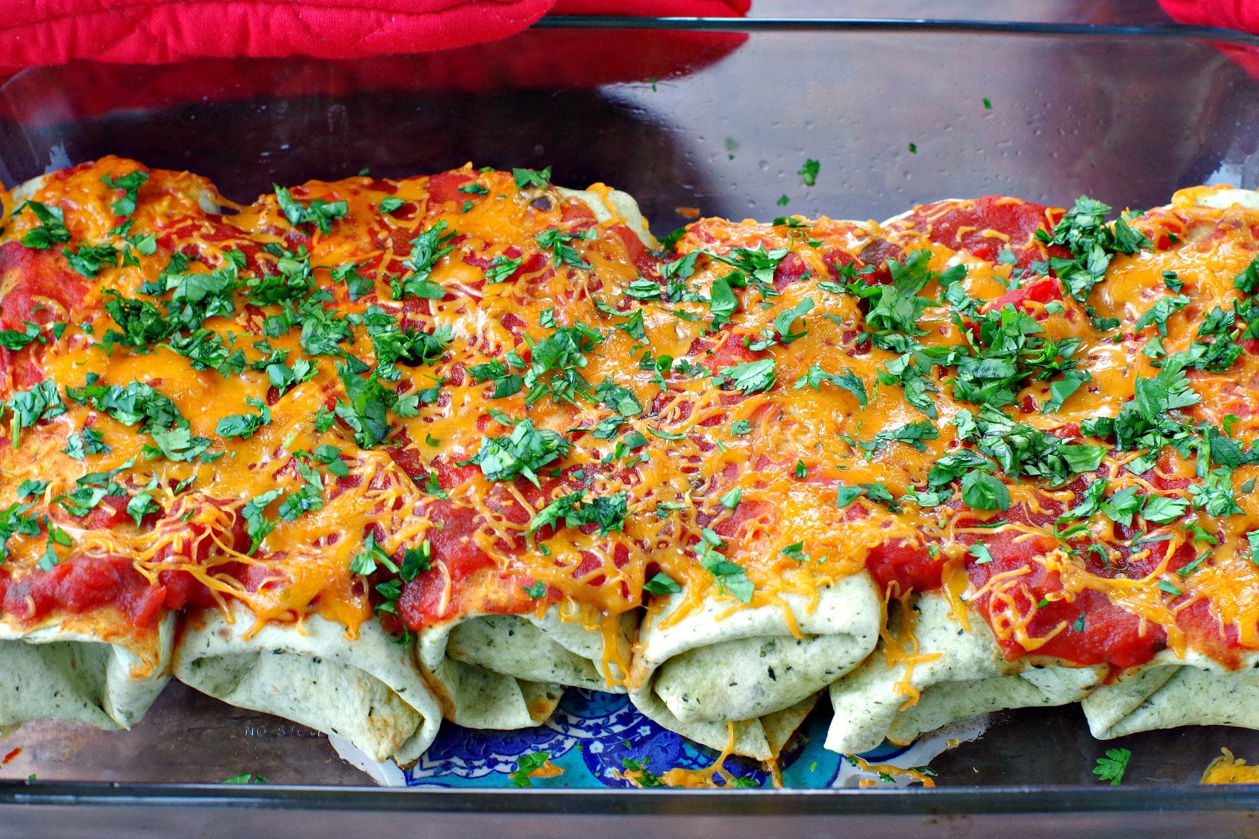 leftover turkey enchiladas with cheese salsa and chopped cilantro sprinkled on in glass casseroled dish with red oven mitts in the background