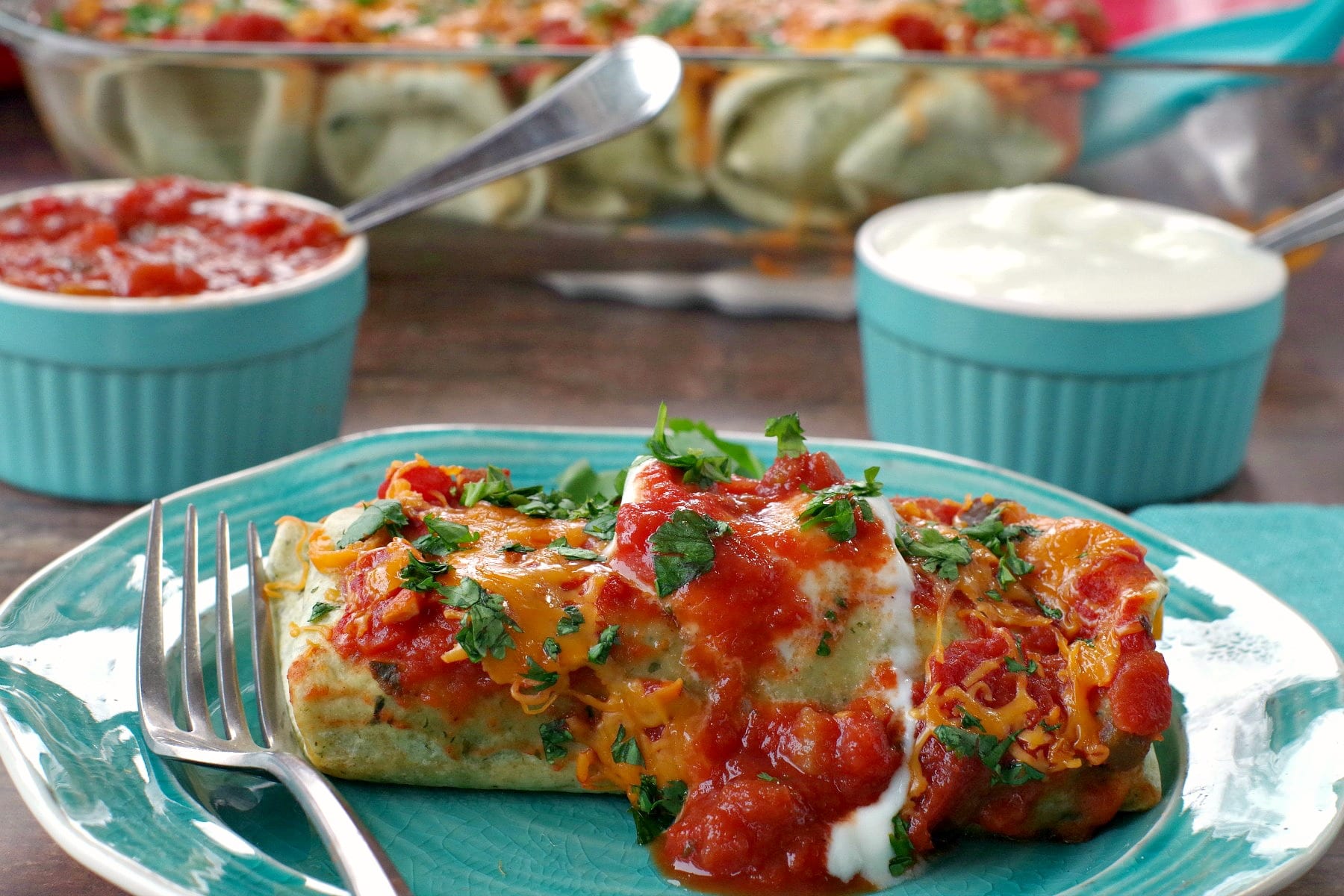 leftover turkey enchilada on a blue plate with glass dish of enchiladas in background and blue ramkins with sour cream and salsa right behind the dish