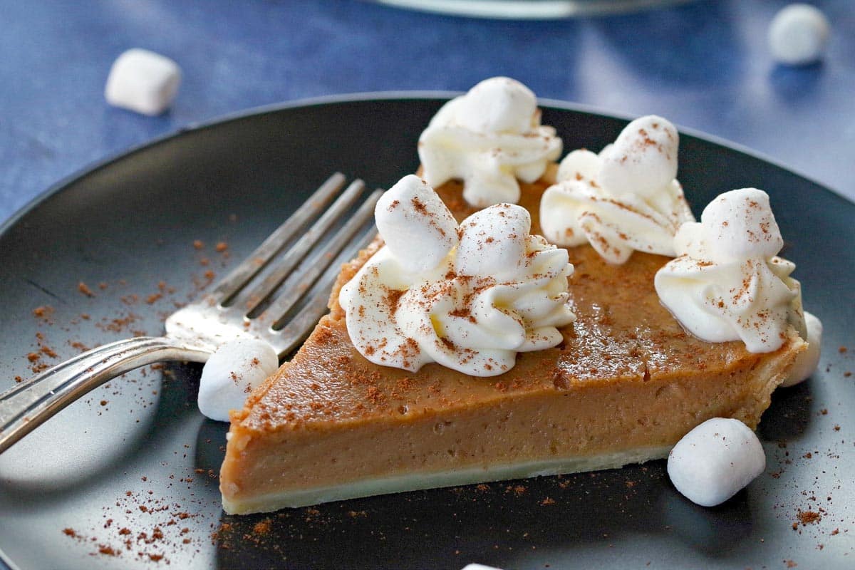 Sweet potato pie on black plate on blue background with marshmallows around and pie in the background.