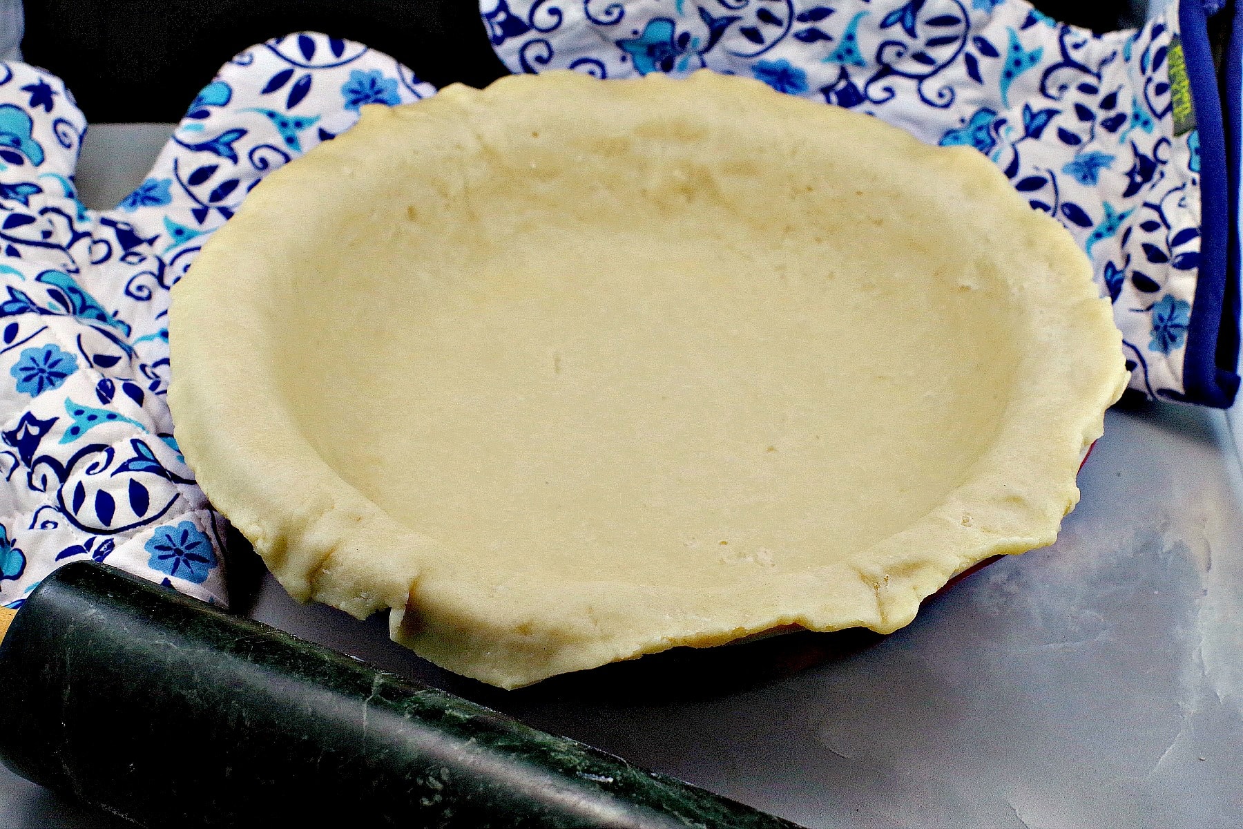 uncooked cold water pastry (no fail pie crust) in pie pan with oven mitts in the background and rolling pin in front