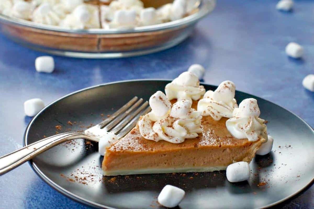 Piece of Sweet Potato Pie with Marshmallow Whipped Cream on Black plate on blue background with whole pie in glass pie plate in the background