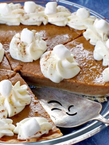 Sweet potato pie with marshmallow whipped cream in glass pie plate with piece missing and pie lifter in missing spot.