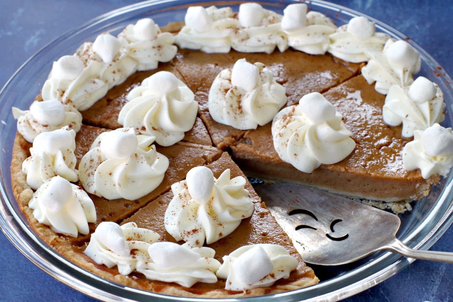 A freshly baked pumpkin pie is topped with dollops of whipped cream and small marshmallows. One slice is missing, and a metal pie server rests on the transparent glass dish, which sits on a dark blue surface.