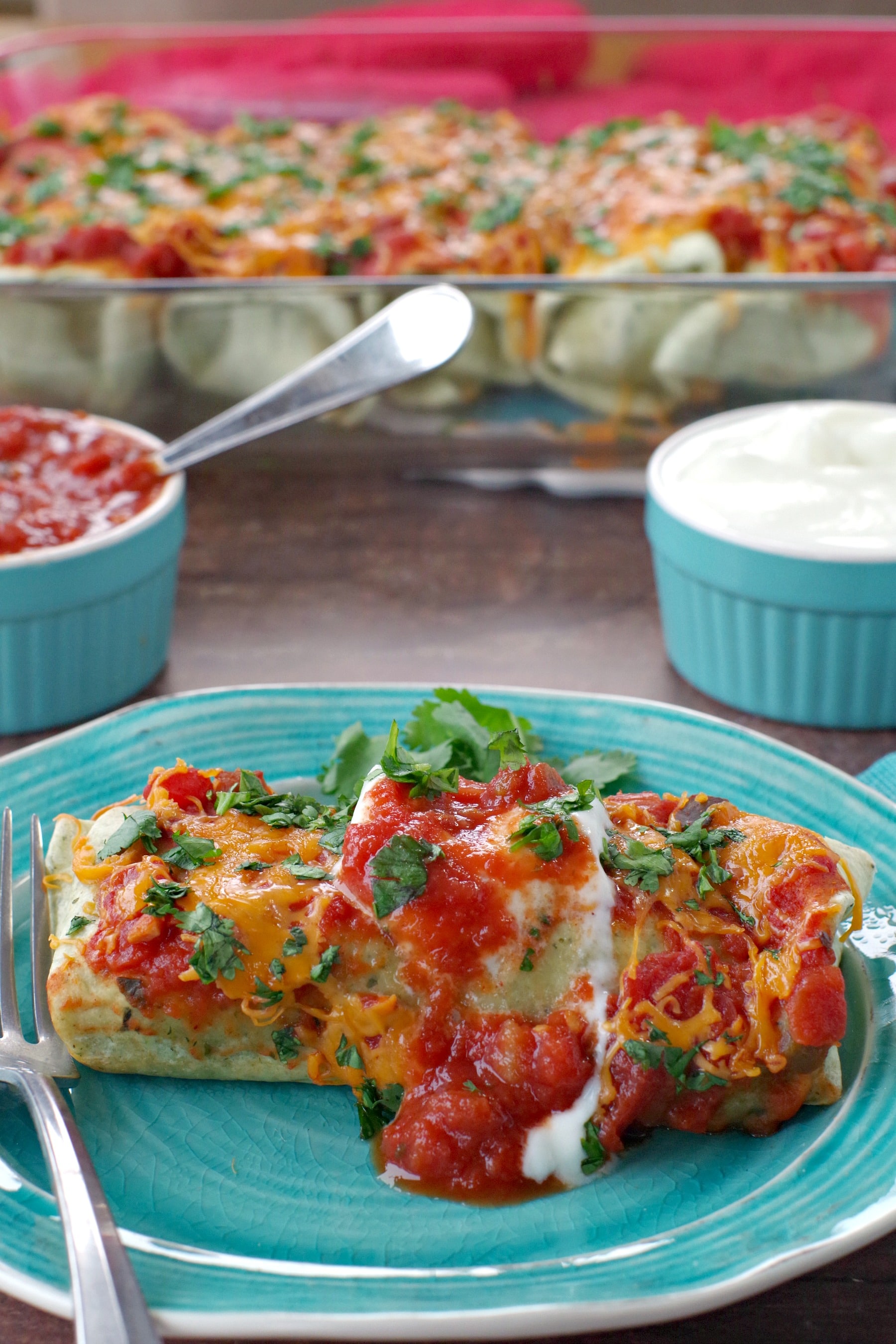 leftover turkey enchilada on a blue plate with glass dish of enchiladas in background and blue ramkins with sour cream and salsa right behind the dish