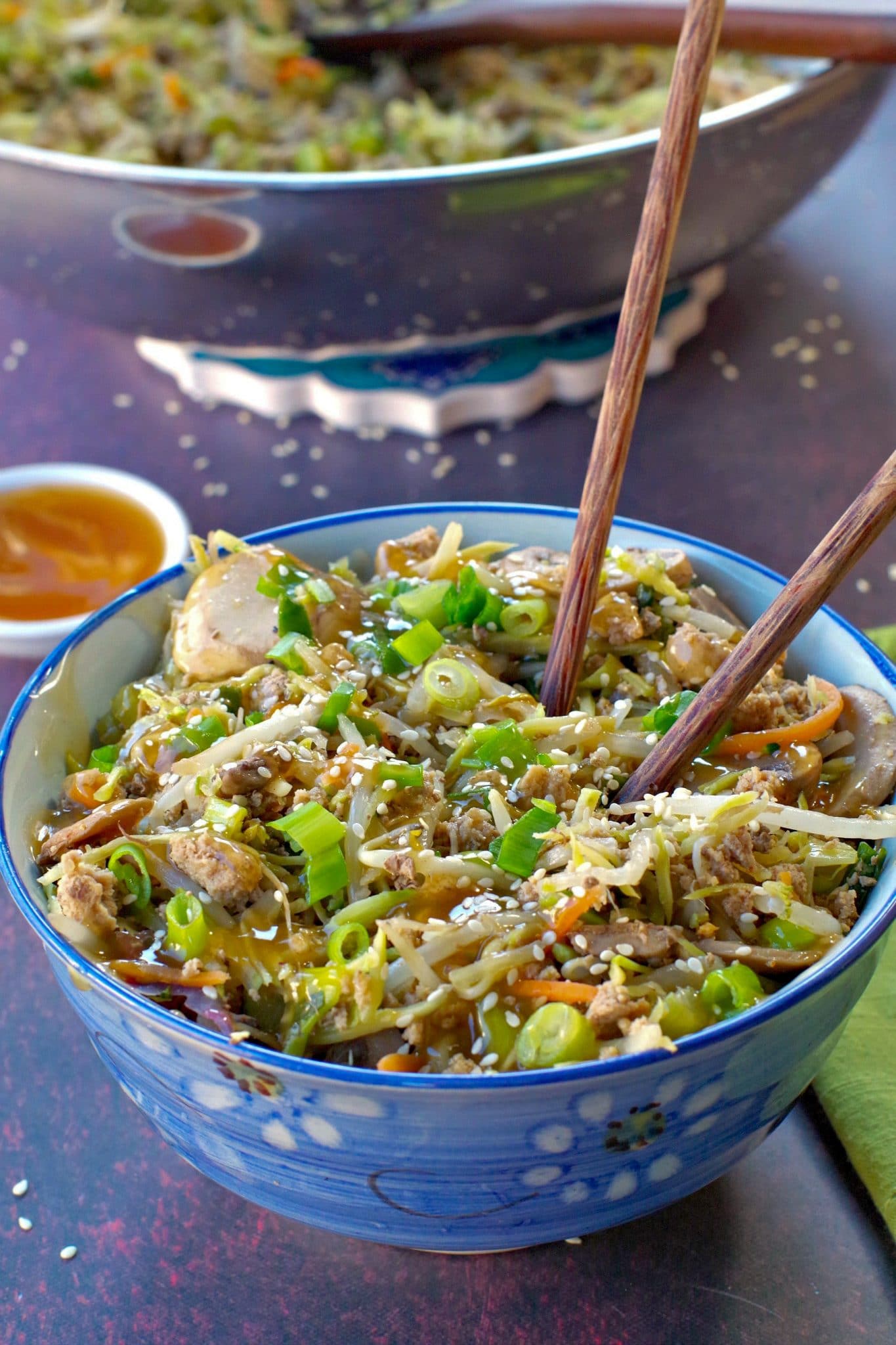 egg roll in a blue flowered bowl with chopsticks and a small white dish of plum sauce in the background and wok with more egg roll in a bowl in the background