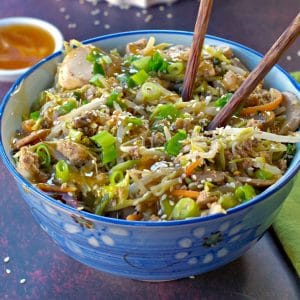 egg roll in a blue flowered bowl with chopsticks and a small white dish of plum sauce in the background and wok with more egg roll in a bowl in the background