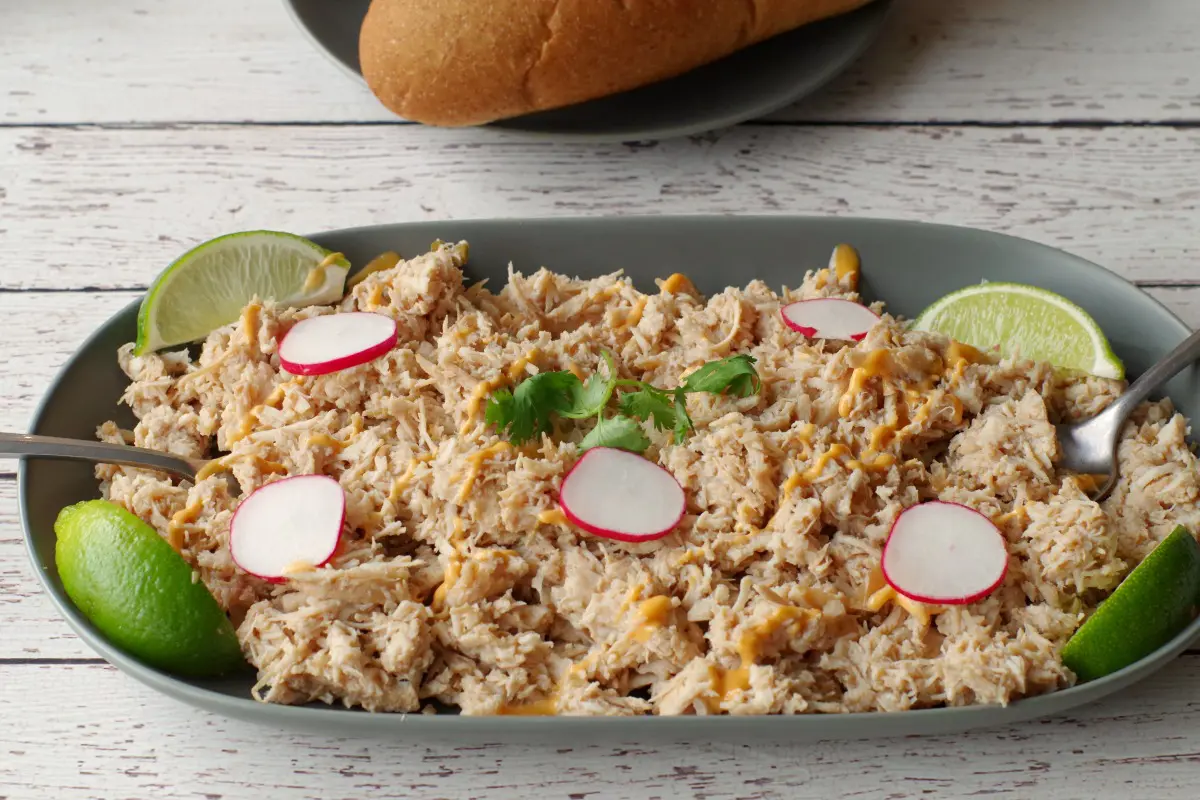 shredded chicken on greyish green plate, garnished with radishes and lime wedges and sub buns in the background on a black plate.