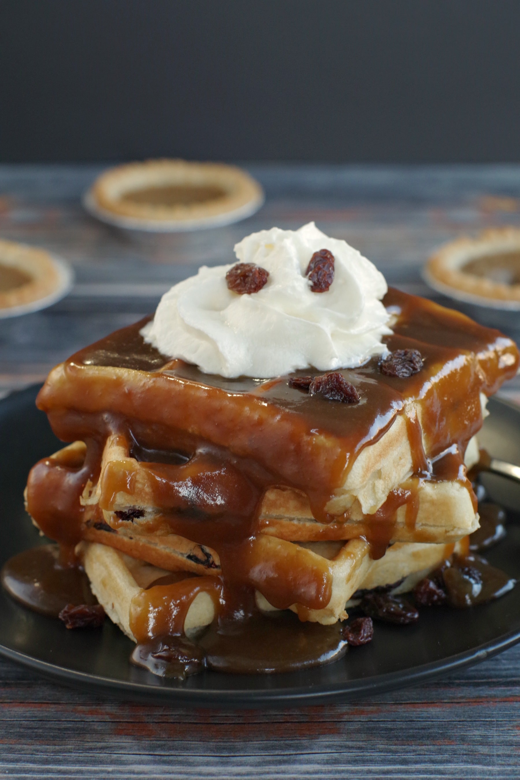 4 Butter Tart Waffles stacked on a black plate (with lots of sauce on them) on a blue wooden background and butter tarts in the background