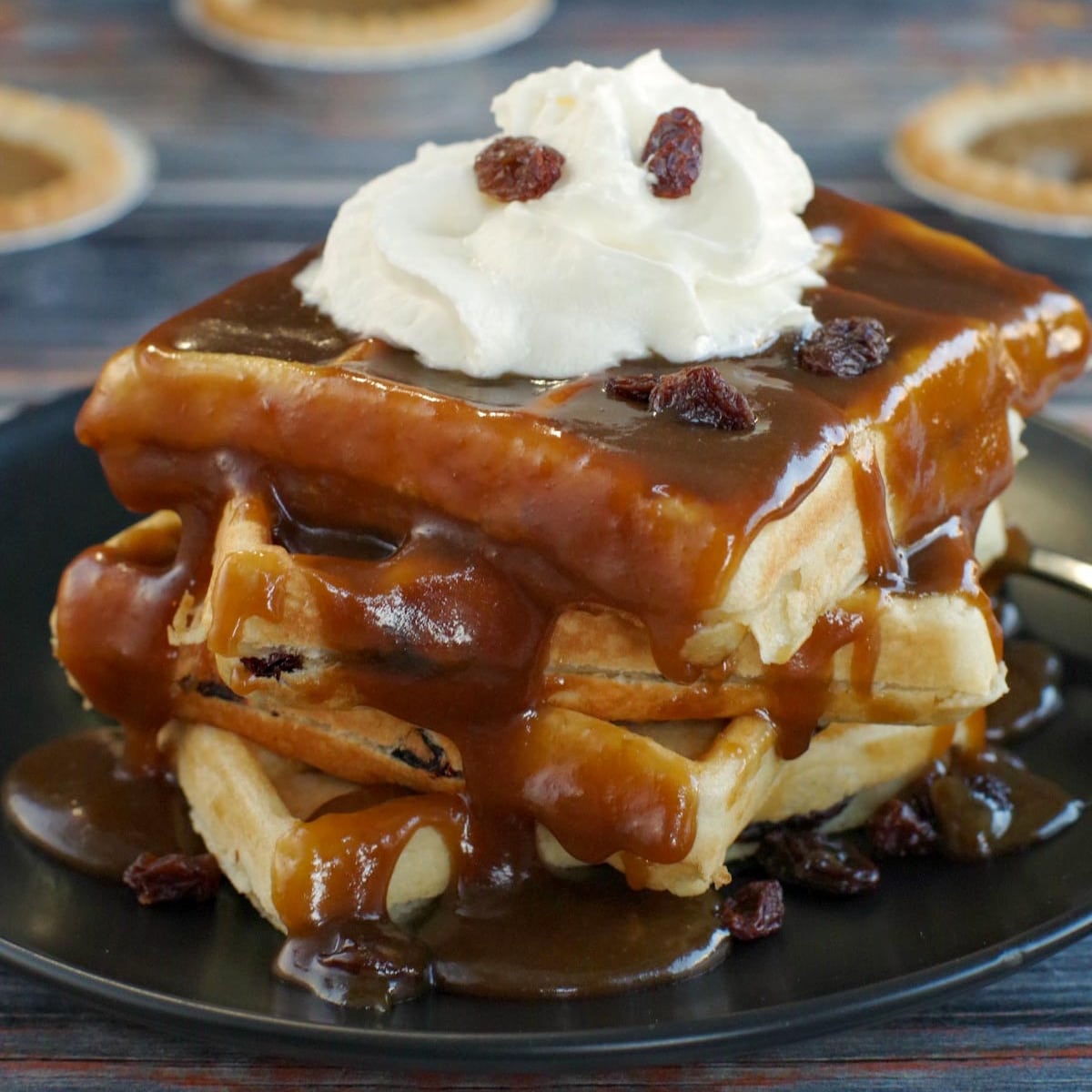 4 butter tart waffles stacked, with butter tart sauce, on a black plate with butter tarts in the backgroun