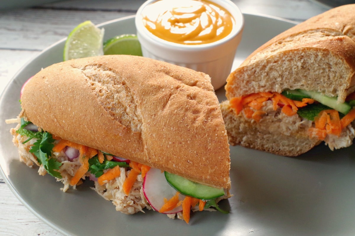 Chicken Vietnamese sub, sliced in half, with a small white dish of sriracha mayo and lime wedges on a greenish-grey plate