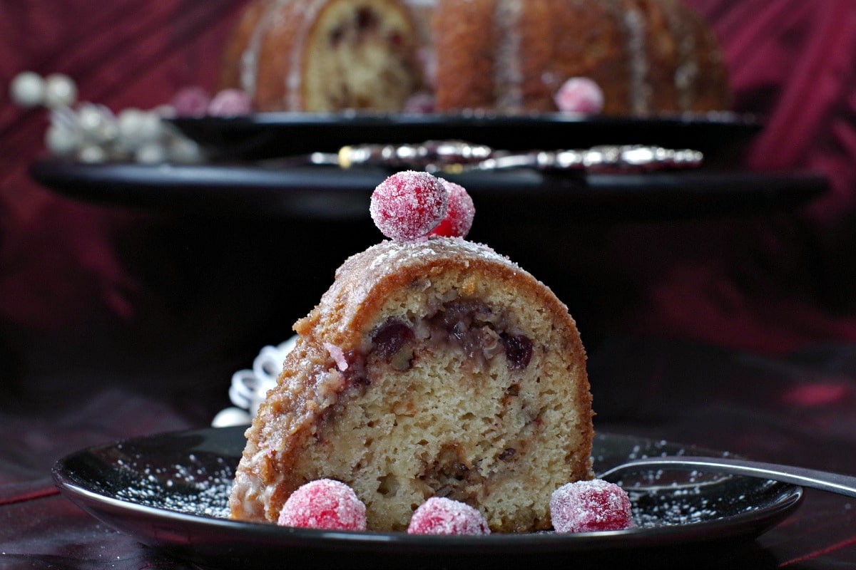 a slice of cranberry cake on a black plate with candied cranberries and a whole cranberry cake in the background