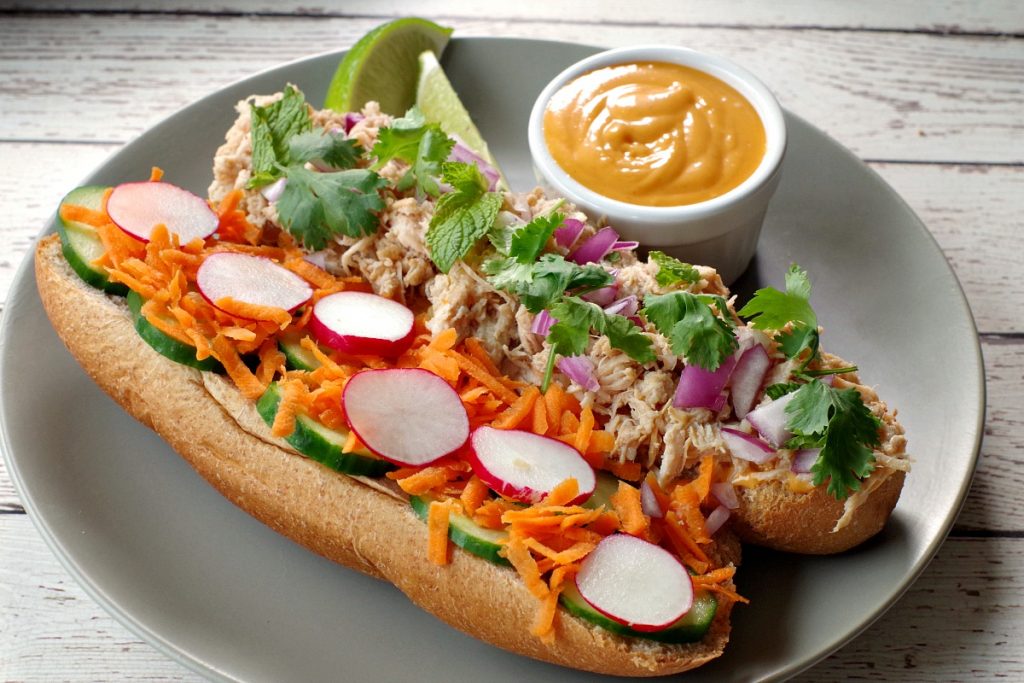 open chicken banh mi vietnamese sub on a greenish grey plate with lime garnish and small white dish of sriracha mayonnaise