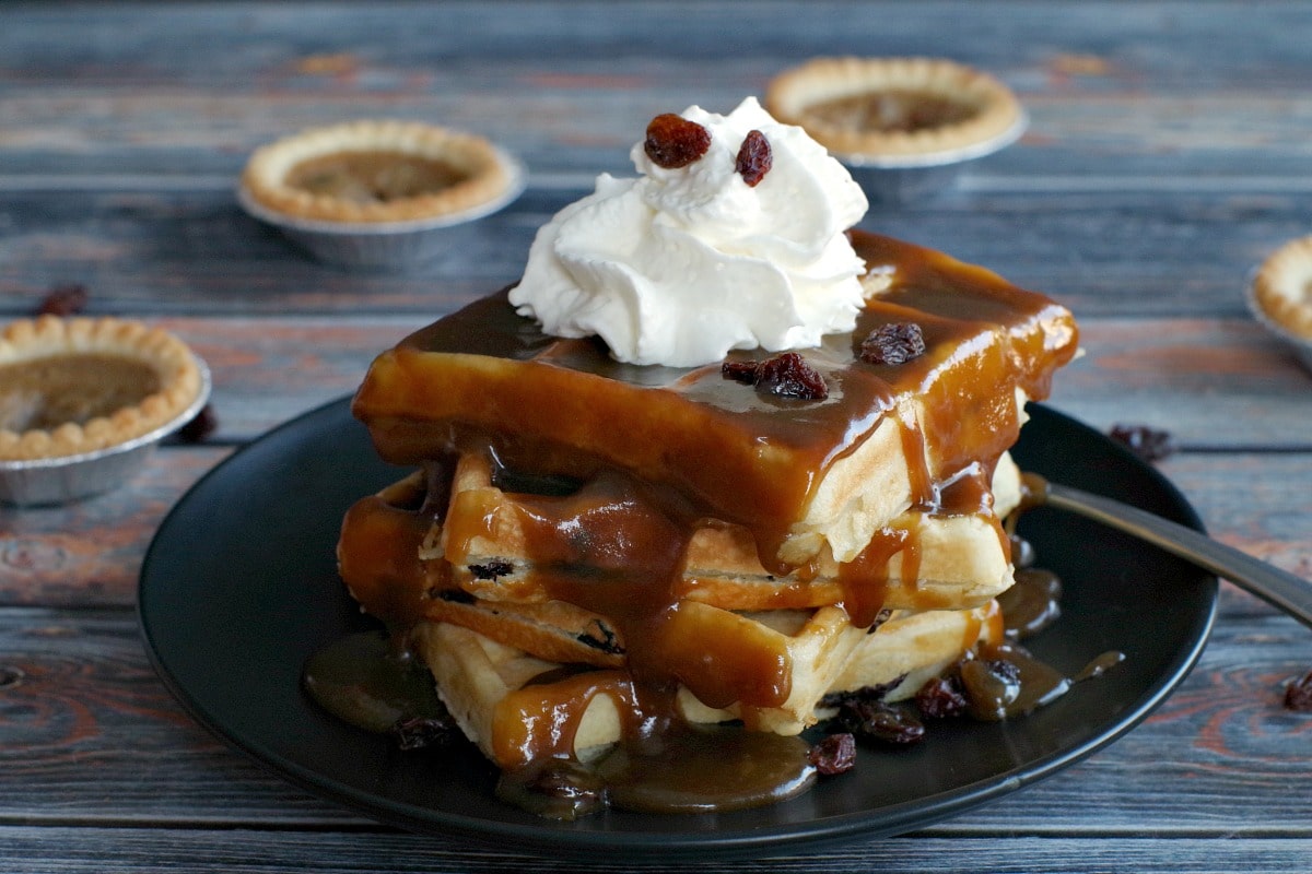 4 Butter Tart Waffles stacked on a black plate (with lots of sauce on them) on a blue wooden background and butter tarts in the background