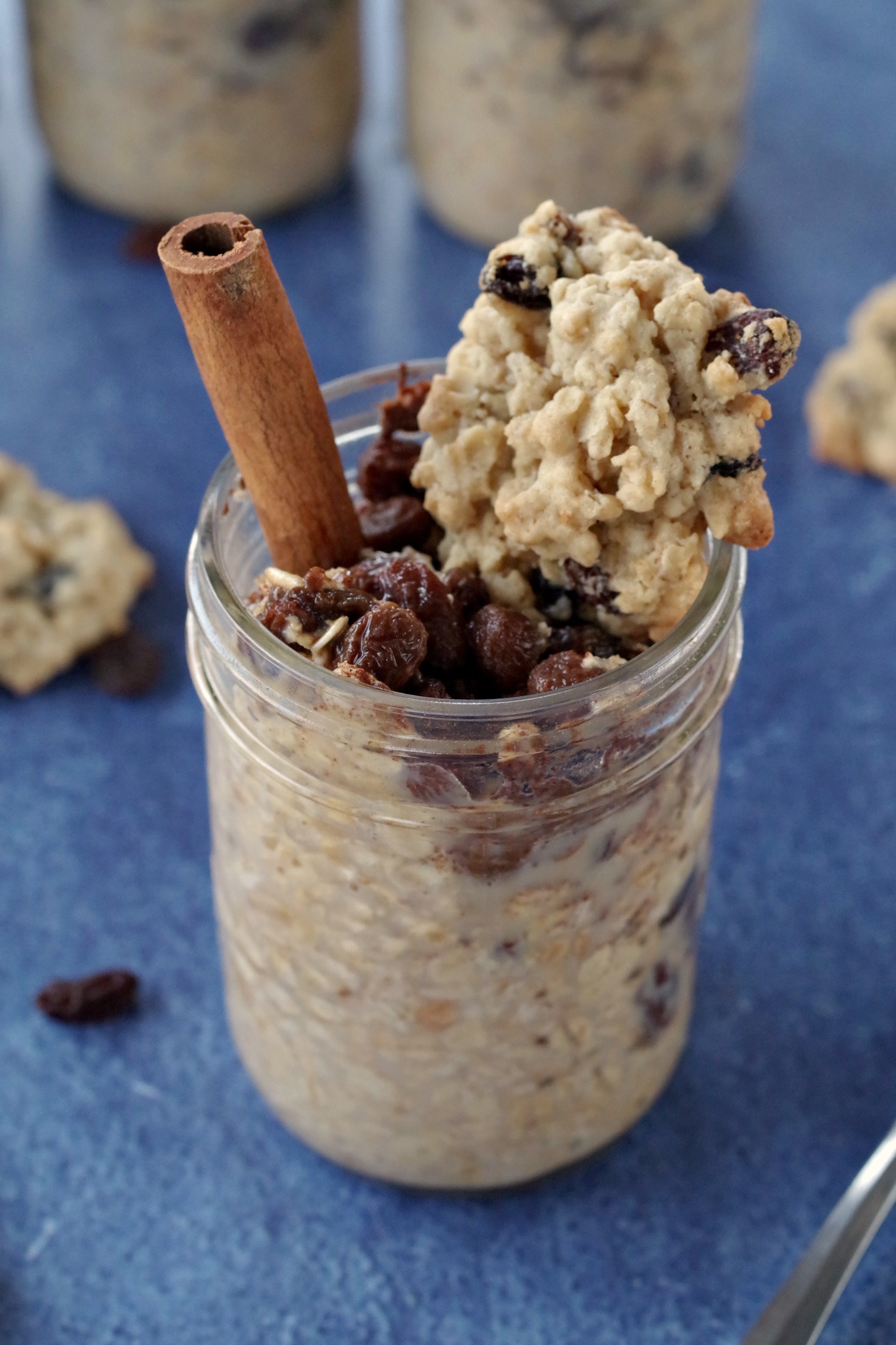 raisin cookie oatmeal in a jar with cookie on top and cinnamon stick on blue background with raisin oatmeal cookies in background