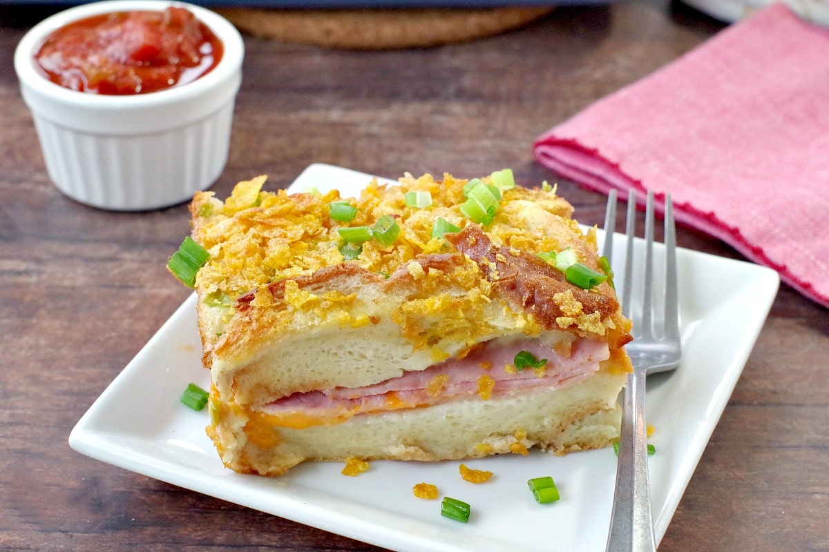 A slice of Christmas morning casserole on a white plate, on brown wooden surface, with fork on red napkin beside it, salsa in a white dish behind