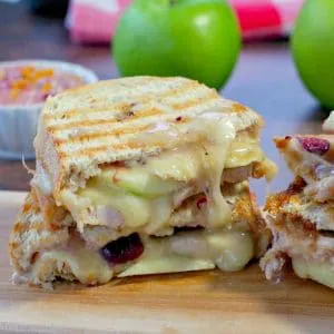 Turkey Panini with cranberry and brie on a cutting board with green apples and cranberry aioli in the background