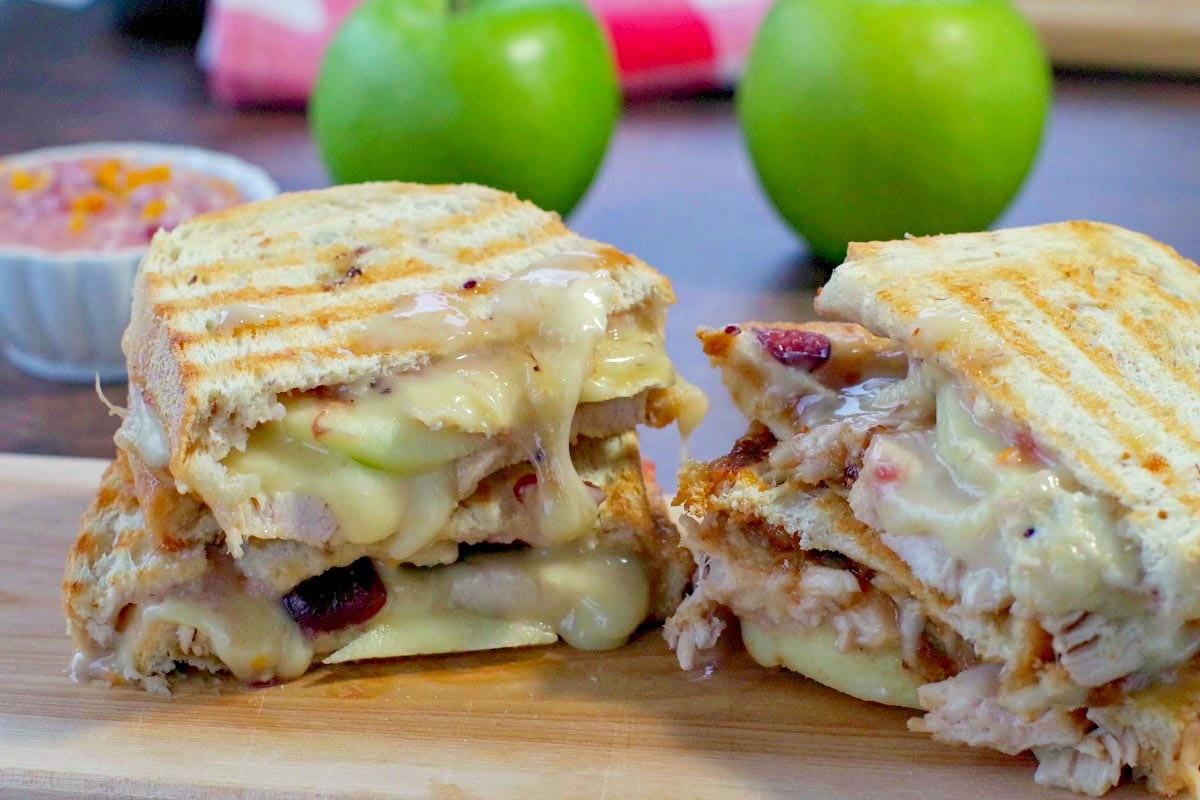 Turkey Panini with cranberry and brie on a cutting board with green apples and cranberry aioli in the background
