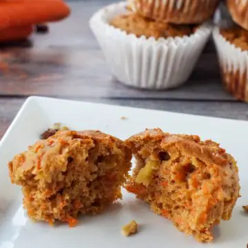 carrot muffin on blue wood-like surface, pulled into 2 pieces , with wrapper, more muffins and carrots in background